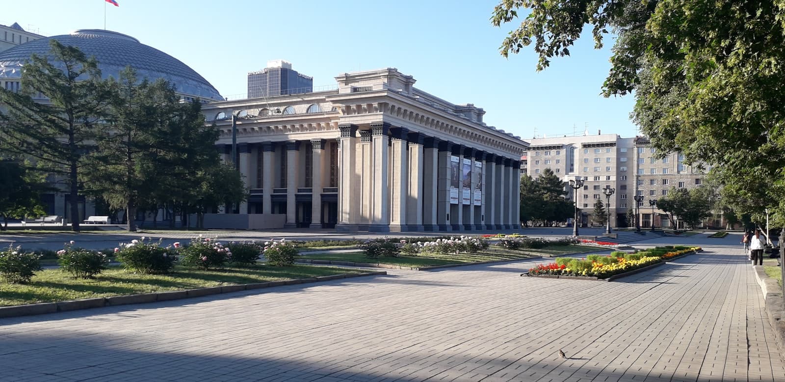 Novosibirsk — few people walking near ancient historic building surrounded with tall and green trees during daytime