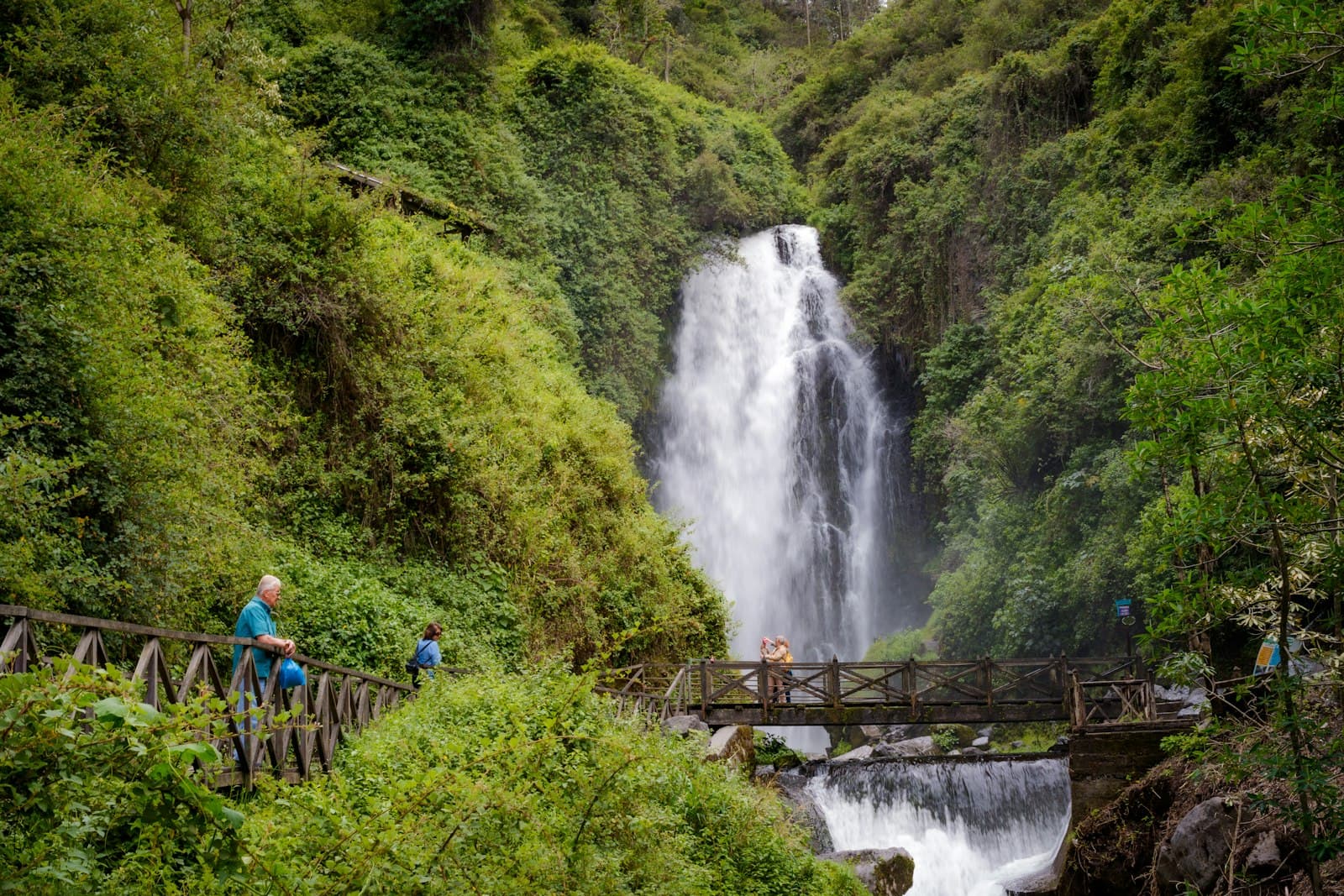 Otavalo — A group of people walking across a bridge next to a waterfall