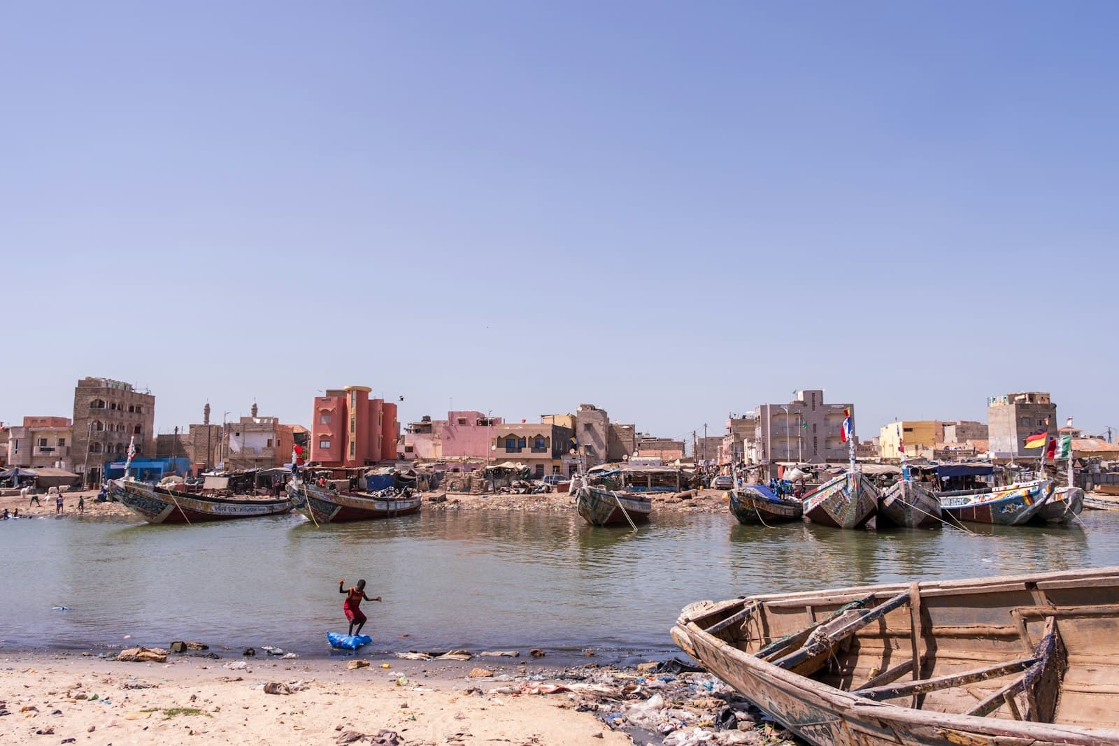 Thiès — Child playing near old boats in a bustling coastal town.