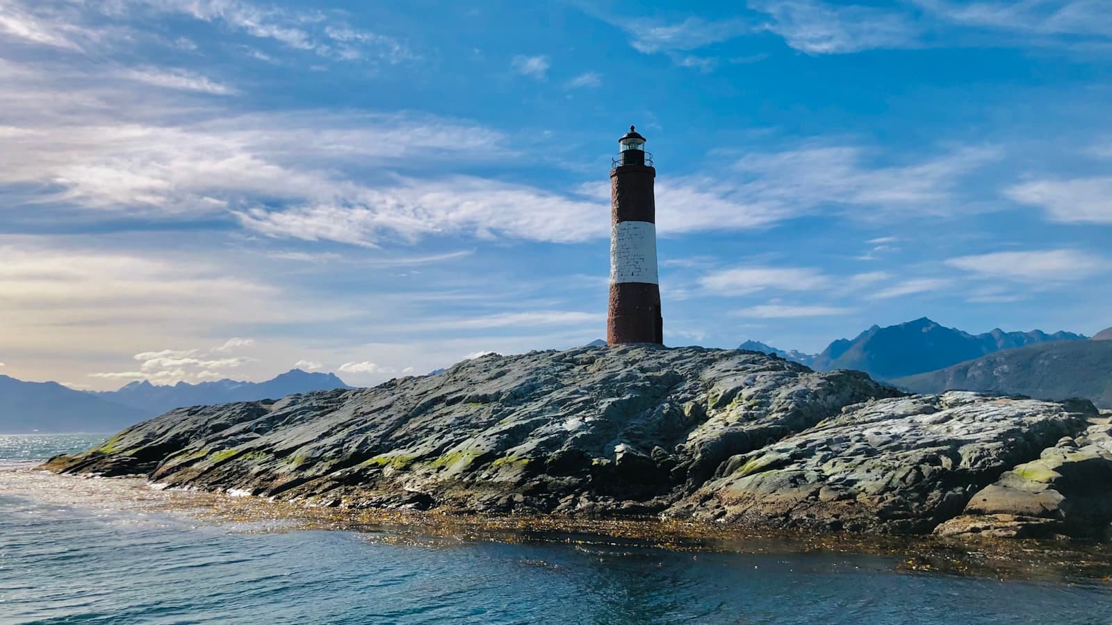 Ushuaia — brown and black lighthouse on rocky hill near body of water under blue and white cloudy